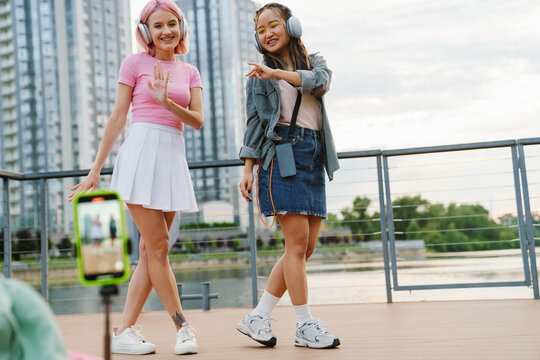 Two cheerful girls making video with smartphone while standing on city waterfront