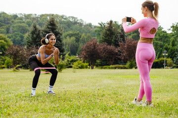 Woman making video with smartphone camera of her friend squatting with resistance band in park