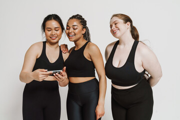 Three cheerful women in sportswear using smartphone isolated over white background