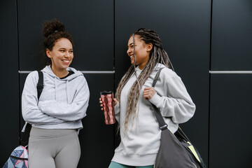 Two cheerful fitness women using smartphone while standing outdoors near black wall