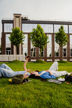 Two female students taking selfie and showing peace sign while laying on grass in campus