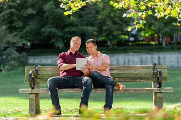 Smiling gay couple, homosexuals sitting on bench on street, using digital tablet, online shopping - obrazy, fototapety, plakaty