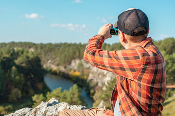 man in cap red plaid shirt sitting in rocks and taking photo on smartphone of mountains autumn...