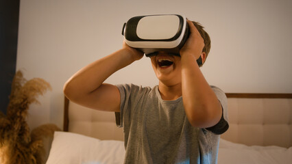 Funny enthusiastic smiling child kid boy schoolboy play video game using innovative digital VR goggles playing online 3d AR technology in metaverse world wearing virtual reality glasses at home on bed