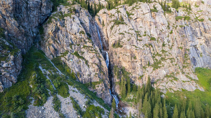 A high waterfall cascades in a mountain gorge. Coniferous trees, Christmas trees and grass grow. The river runs. Hiking trails. Top view from a drone. Sunset. Burkhan Bulak Waterfall, Kazakhstan