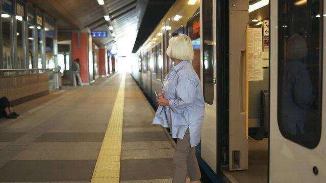 Happy senior 50s caucasian woman go out from train. Blonde wears denim jacket. People on public transport platform. High quality FullHD footage