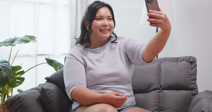 A chubby Asian woman using smart phone to take selfie while sitting on sofa at home.