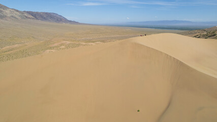 A large sand dune in the middle of the steppe. Drone view of a huge pile of sand. Tourists are walking, enjoying the view. A river runs in the distance and grass grows. The sky with white clouds