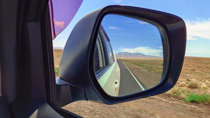 View from the car interior in the side mirror. The car is driving along the road in the middle of the steppe. Dry bushes, poles. In the reflection of a white car, a road and mountains with the sky