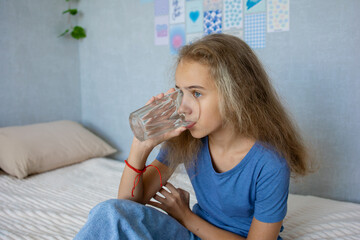 A cute teenage girl drinks water from a clear glass while sitting on the bed in her bedroom....