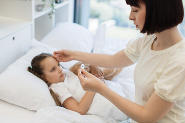 Caucasian dark haired woman sitting on white bed, looking on digital thermometer and holding hand on child forehead. Focused caring mother controlling temperature of her sick daughter at home.