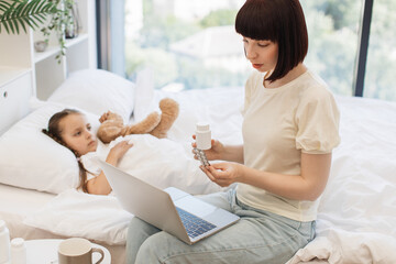 Middle aged woman having consultation with pediatrician on taking pills while caring little sick daughter at home. Loving mother sitting on bed near sad cute child and holding modern laptop on knees.