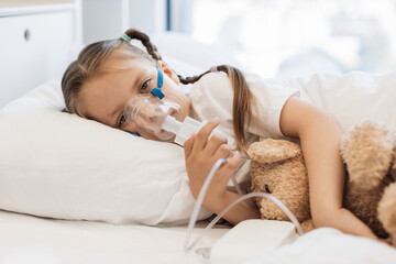 Portrait of little sad child lying in white bed and hugging bear toy while inhaling medication...