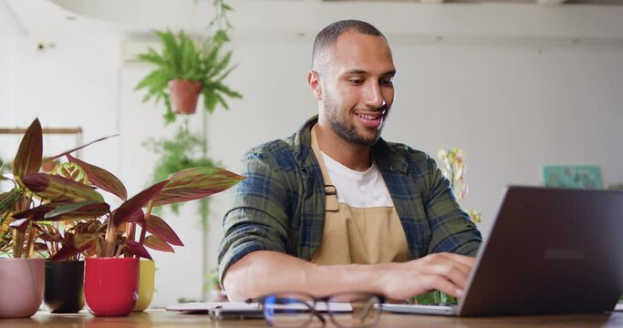 Owner male of flower shop working on laptop computer in workplace sitting in floral store. Man florist in apron looking at laptop and making sales order.