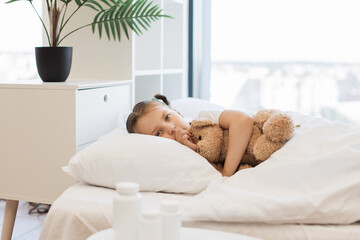 Fatigued cute girl with influenza coughing in clenched fist while lying on bed with soft white linen. Little blond child finding comfort in embrace of her teddy bear toy.
