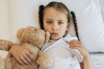 Top view of sick little girl hugging her beloved bear toy while lying on bed with digital...