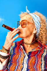 Portrait of senior woman in stylish accessories and sunglasses smoking cigar over blue studio...