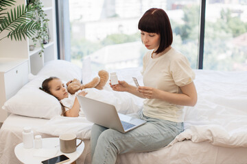 Middle aged woman having consultation with pediatrician on taking pills while caring little sick daughter at home. Loving mother sitting on bed near sad cute child and holding modern laptop on knees.