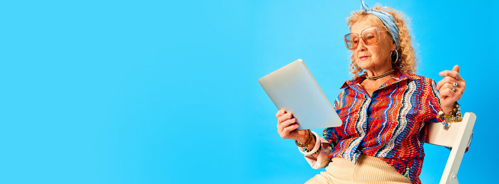 Senior elegant woman in stylish clothes sitting on stairs and reading news on tablet over blue studio background. Concept of human emotions, fashion, elderly people, lifestyle, creativity. Ad