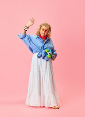 Hipster vibe. Senior beautiful woman in stylish casual clothes, shirt, skirt and sunglasses posing with skateboard over pink background. Concept of emotions, fashion, elderly people, lifestyle. Ad