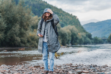 A young woman traveler in a raincoat using smartphone on the bank of a mountain river. Hiking and...