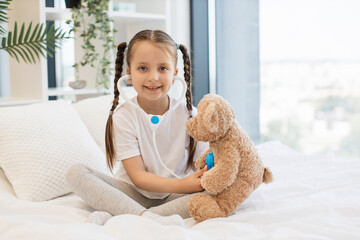 Little caucasian girl listening heartbeat of teddy bear with toy stethoscope in cozy bed against background of panoramic window with city view. Concept of childhood, game and treatment.