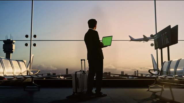 Full Body Back View Of Asian Businessman With Rolling Suitcase In Boarding Lounge At The Airport, Using Green Screen Laptop While Waiting For Flight, Airplane Takes Off Outside The Window
