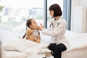 Competent female doctor conducting detailed examination of adorable little girl sitting on cozy bed and hugging teddy bear. Medical worker checking lymph nodes of sick patient during domestic visit.