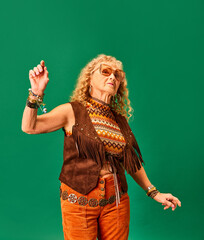 Hippie outlook. Youthful senior woman in stylish clothes, accessories and sunglasses dancing over green studio background. Concept of human emotions, fashion, elderly people, lifestyle, creativity. Ad