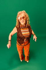 Hippie outlook. Youthful senior woman in stylish clothes, accessories and sunglasses dancing over green studio background. Concept of human emotions, fashion, elderly people, lifestyle, creativity. Ad