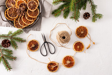 Top view of white textured background with homemade garland of dried oranges, spruce branches, orange slices and craft scissors. Flat lay. decoration.