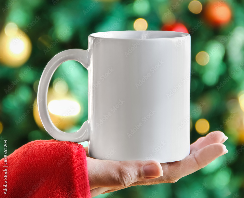 Wall mural Girl is holding white mug in hands with green christmas tree,  bokeh lights, red sweater  and blur tree decoration. Blank 11 oz white xmas ceramic cup	