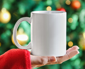 Girl is holding white mug in hands with green christmas tree,  bokeh lights, red sweater  and blur...
