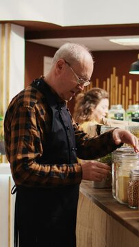 Vertical video Portrait of elderly vendor working in zero waste organic food supermarket, adding pantry staples on shelves. Older employee restocks local grocery store with bulk products in
