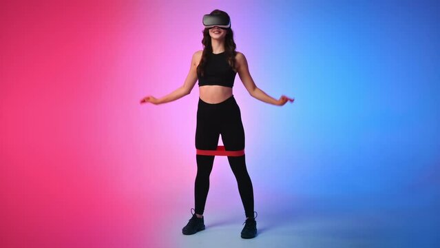 Young smiling woman in a tracksuit in virtual reality VR headset squatting with elastic band in a studio with blue and red background