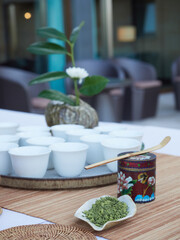 Matcha tea powder on leaf-shaped bowl, small pot decorated and white bowls on tray. Out of focus vase with green leaves and white flower.