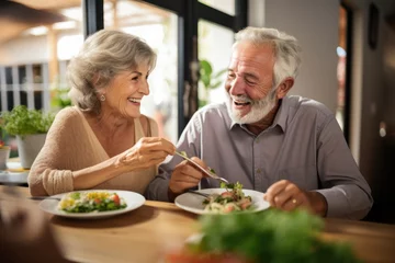 Foto op Canvas Food, retirement and a senior couple in an assisted living home while eating a meal for nutrition. Cute, love or smile with a happy elderly man feeding his wife in the dining room of a house © Tisha