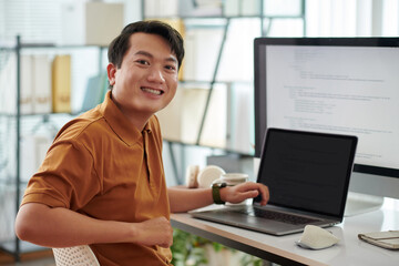 Portrait of cheerful senior developer sitting at desk with computer and laptop with programming...