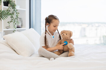 Adorable caucasian girl dressed in domestic clothes using toy stethoscope to checking respiratory system of small teddy bear. Cute doctor with braids hairstyles spending time with pleasure at home.