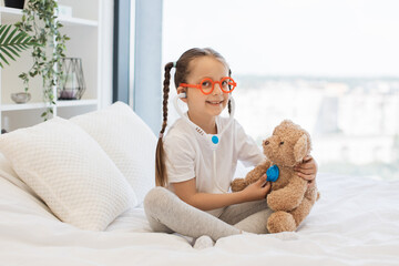 Portrait of cheerful caucasian girl in funny glasses spending time with toy doctor set to treating...