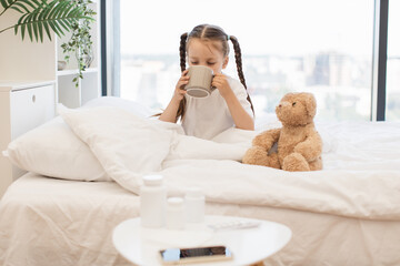 Tired caucasian girl taking sip of hot tea to easing sore throat while warming herself in cozy bed....
