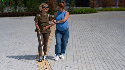 Caucasian pregnant woman leading blind elderly lady outdoors.