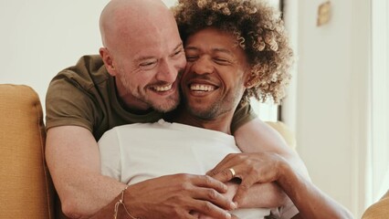 Diverse gay couple at home, sharing love and tenderness on the sofa. A black male with an afro hairstyle smiles, feeling deeply in love with his Caucasian partner, and they share laughter - Powered by Adobe