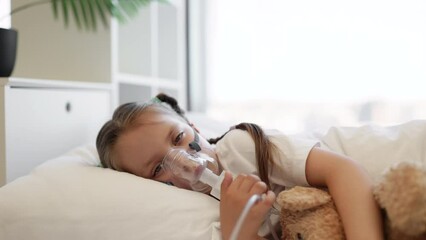 Portrait of little sad child lying in white bed and hugging bear toy while inhaling medication through nebulizer face mask. Upset caucasian girl resting and treating in bright bedroom.