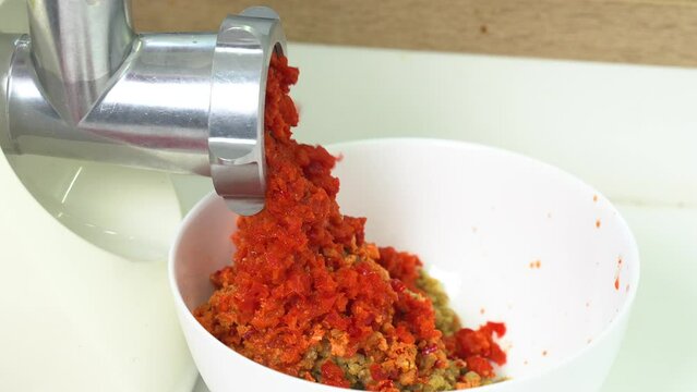 Grinding red pepper in a meat grinder. The resulting product exits through the holes into the plate. Close-up. Side view.