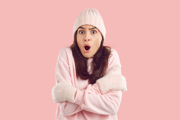 Woman wearing warm light pink hat, sweatshirt and mittens shocked by unexpected extremely low winter temperature feeling very cold, shivering, looking at camera with funny face expression. Studio shot