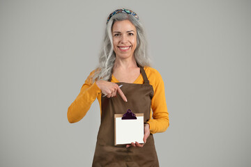 Cheerful senior european lady gray hair framer in apron point finger at clipboard