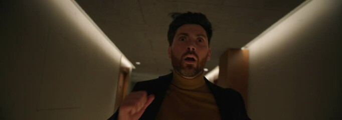 Footage of scared young bearded man in formal clothes running down hallway indoors. Shot of frightened guy in hotel at night. Shot with anamorphic lens. Cinematography technique