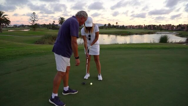 Older man giving golf lessons to an older woman on a golf course. She takes a shot at the ball