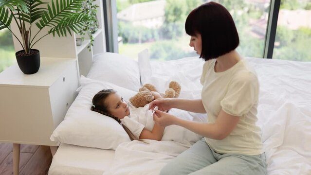 Side view of caucasian woman using digital electronic thermometer for checking temperature of sick daughter. Little kid embracing with teddy bear and enjoying mother's care at home.